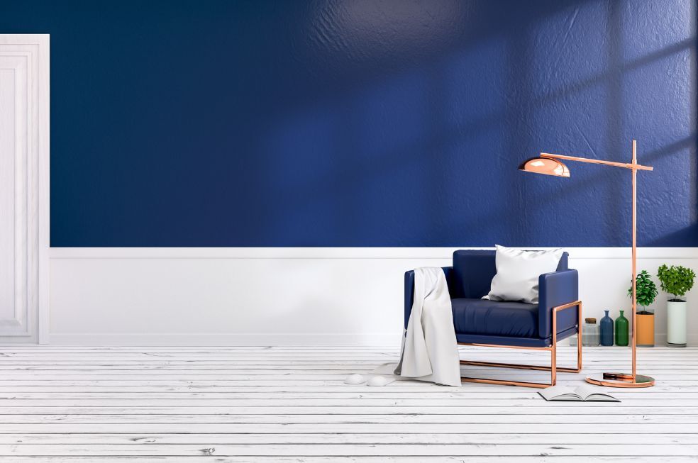 granatowa ściana z białym wykończeniem od dołu i białą podłogą z desek, granatowy fotel z miedzianą lampą